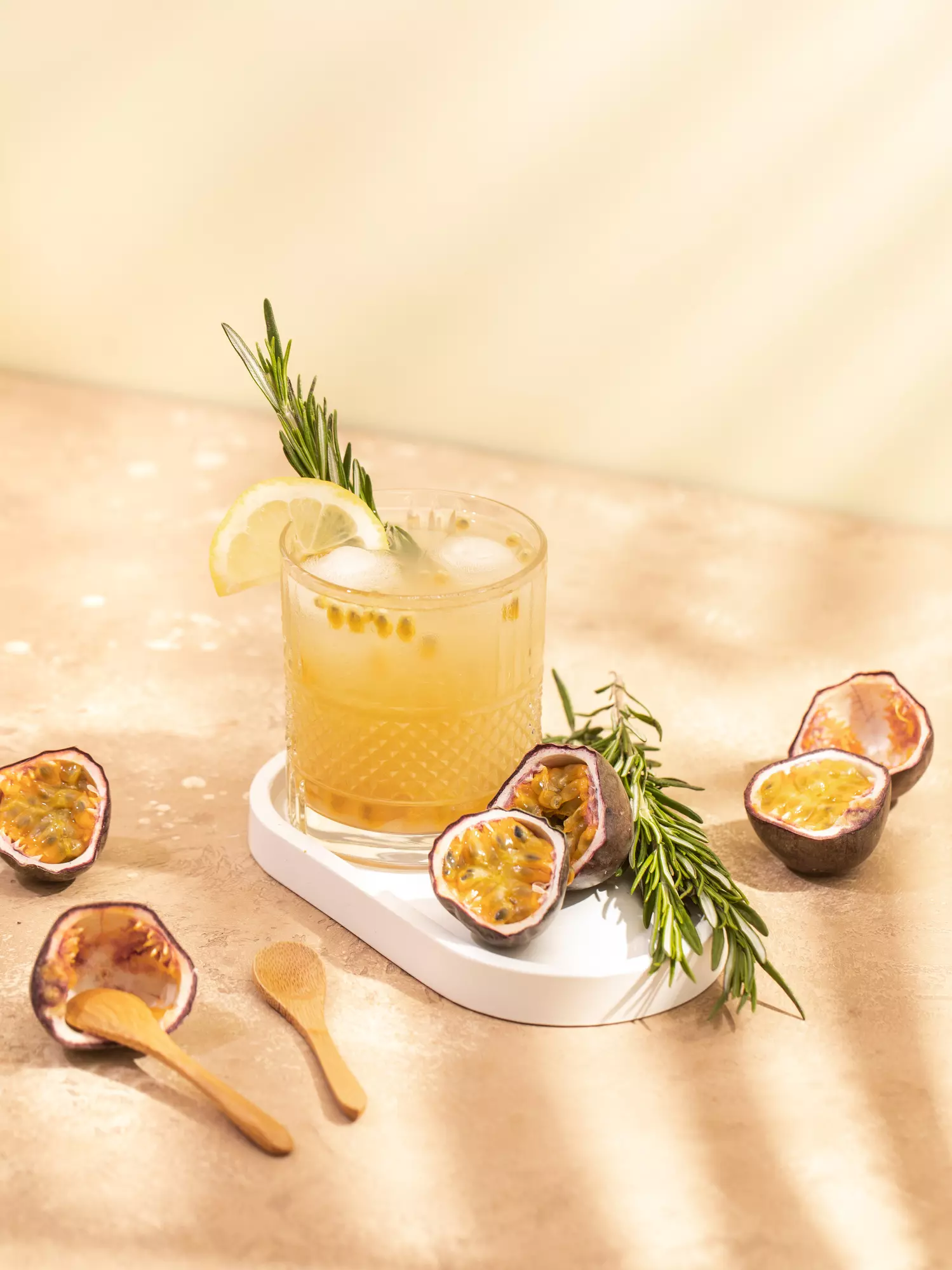 A glass of yellow passion fruit drink with ice, garnished with a lemon slice and rosemary, sits on a tray beside halved passion fruits and wooden spoons.