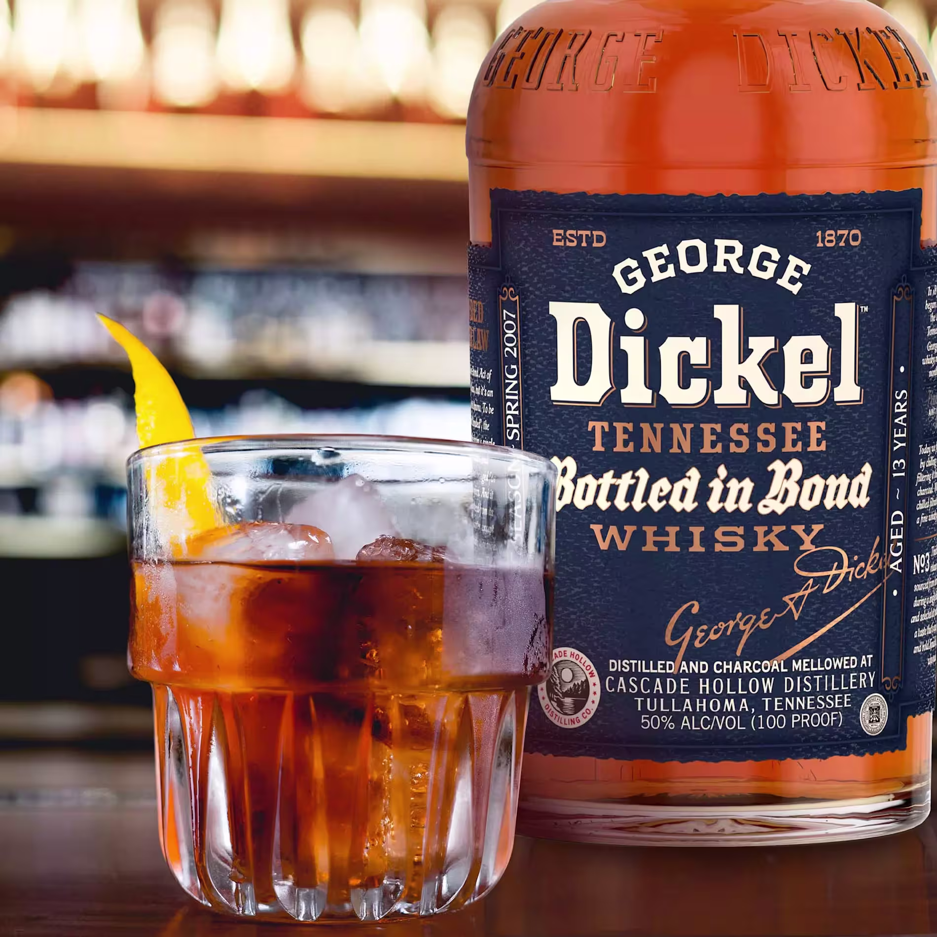 A glass of whiskey with ice and an orange peel garnish sits in front of a bottle of George Dickel Tennessee Bottled in Bond Whisky.