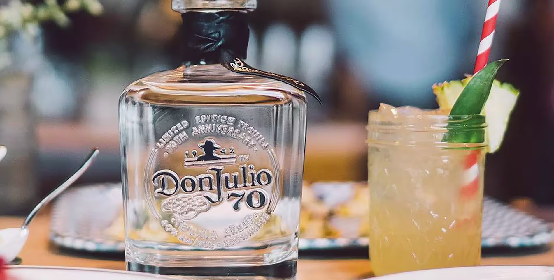 A bottle of Don Julio 70 tequila sits on a table next to a cocktail garnished with a pineapple slice and a red-and-white striped straw.