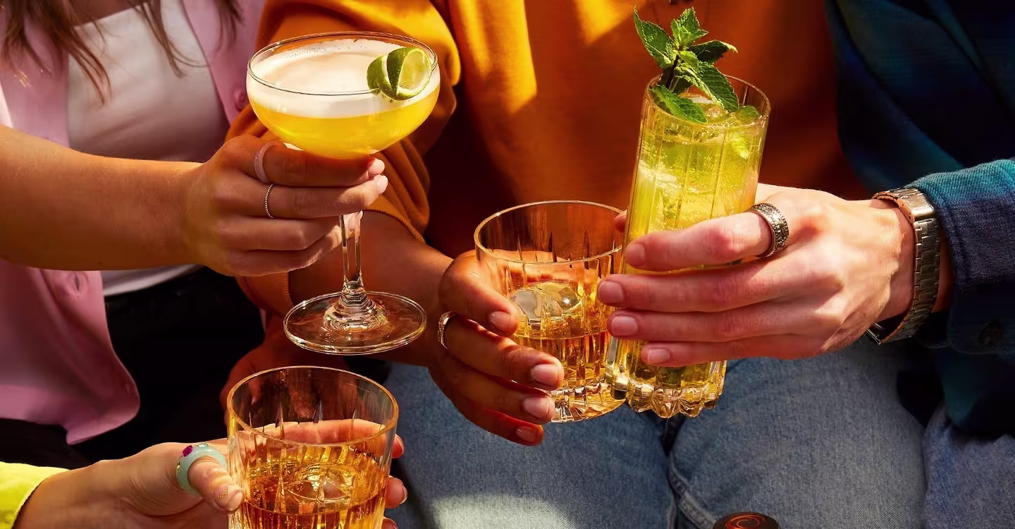 Four people holding various cocktails and glasses in a celebratory toast, with their hands visible and colorful clothing in the background.