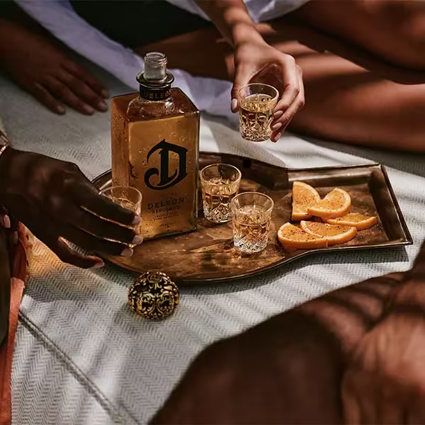 A tray with a bottle of DeLeón tequila, four filled shot glasses, and sliced oranges is surrounded by people reaching for drinks.