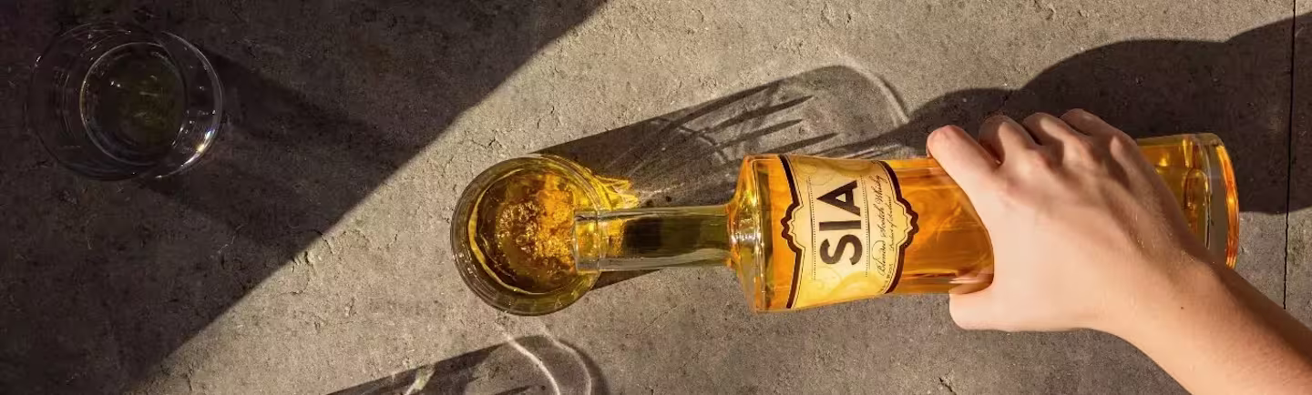 A hand pours SIA Scotch Whisky from a bottle into a glass on a sunlit stone surface.