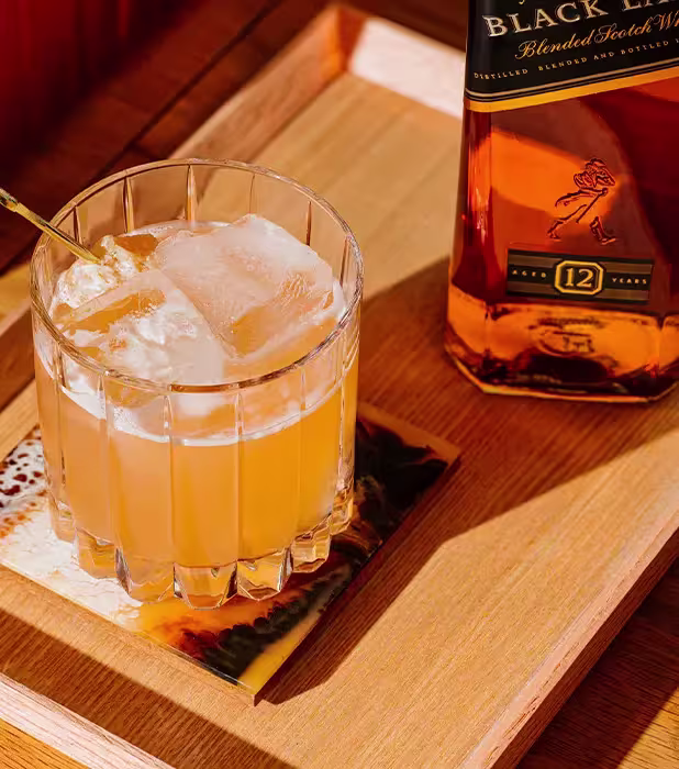 A glass of whiskey with ice and a stirrer sits on a wooden tray next to a bottle of 12-year-old blended Scotch whisky.