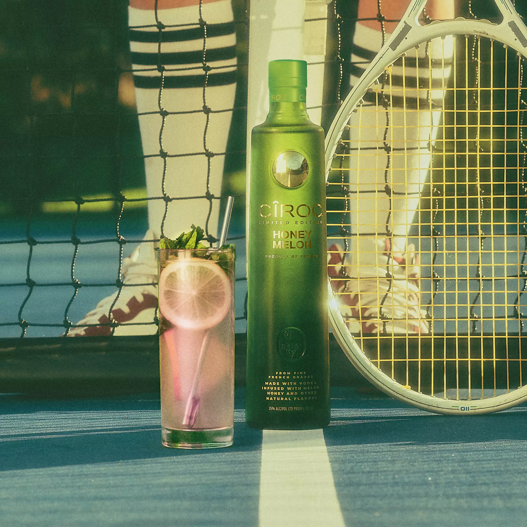 A bottle of Cîroc Honey Melon vodka, a cocktail with lemon and mint, and a tennis racket on a tennis court with a person in striped socks standing behind the net.