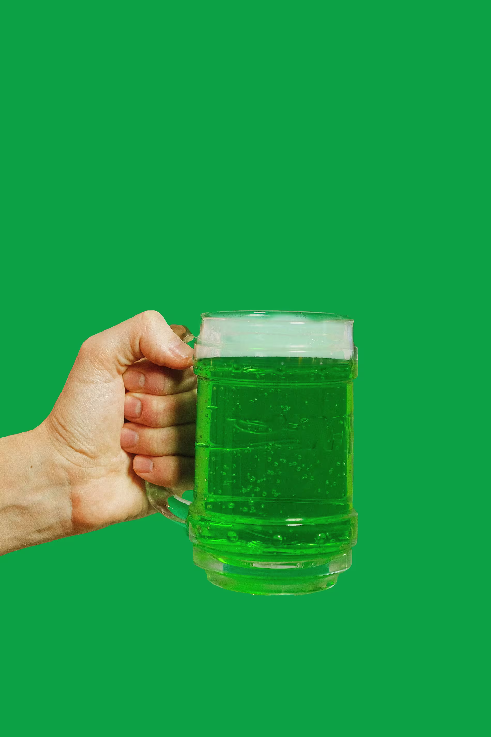 A hand holding a glass mug filled with green beer against a solid green background.