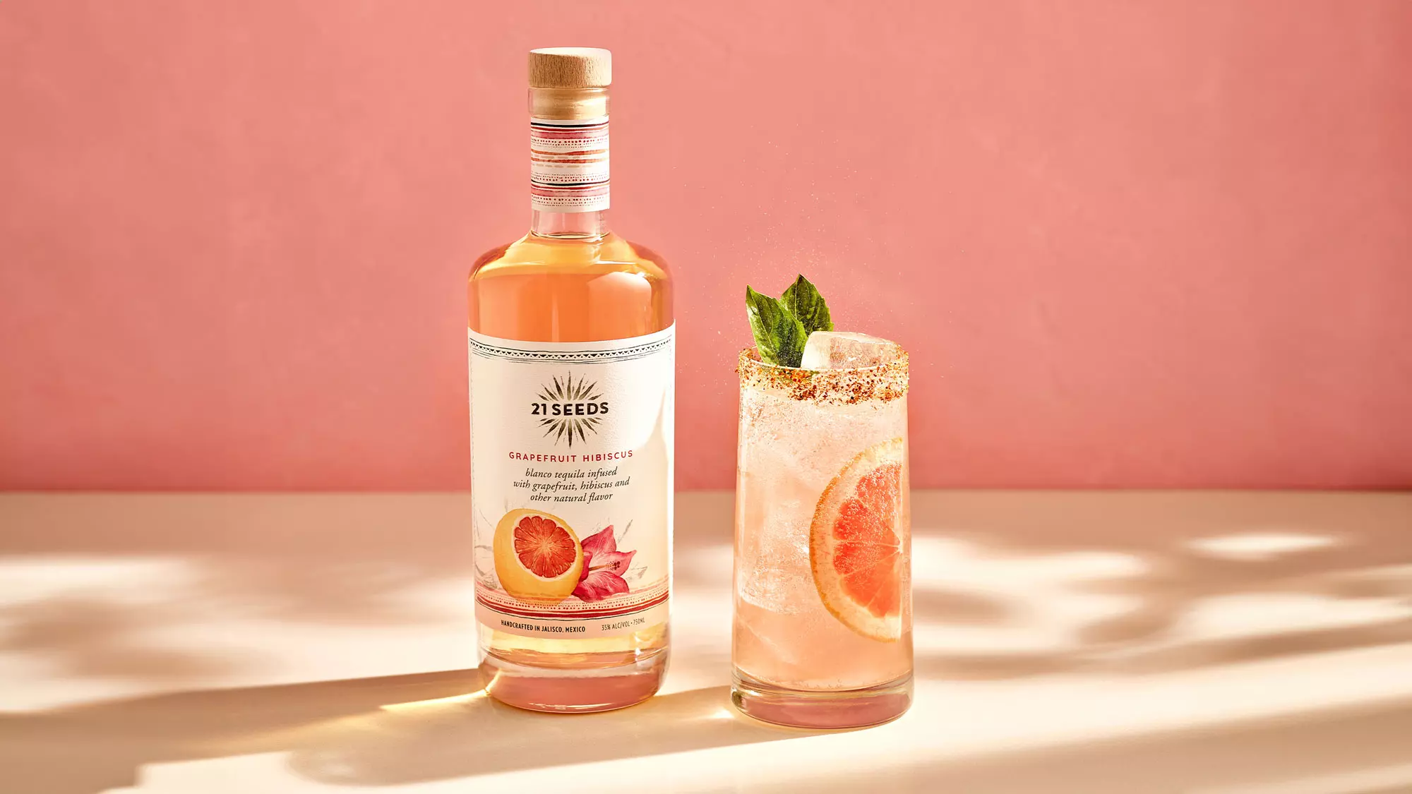 A bottle of 21 Seeds Grapefruit Hibiscus tequila stands next to a cocktail garnished with a grapefruit slice and mint, against a pink background.