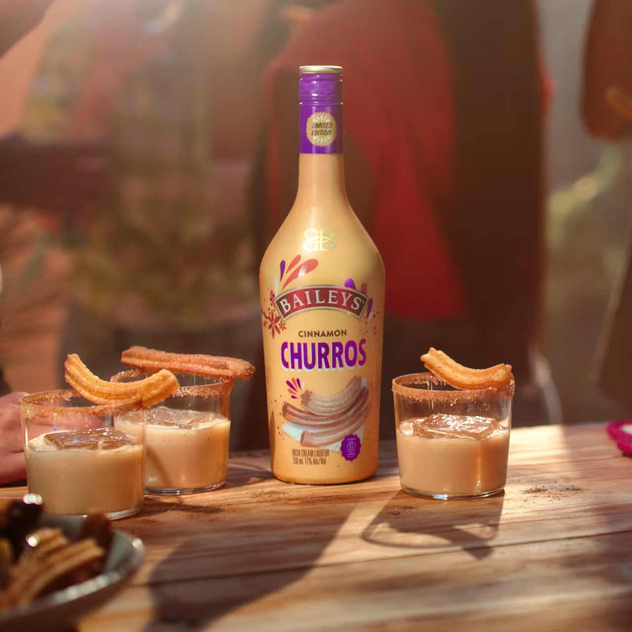 A bottle of Baileys Cinnamon Churros is on a wooden table with three glasses of the drink, each garnished with a churro. People are blurred in the background.
