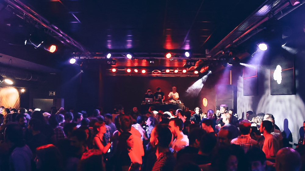 Sala X Sevilla llena de energía con luces vibrantes y un DJ animando la noche mientras el público disfruta de la música y el ambiente festivo.