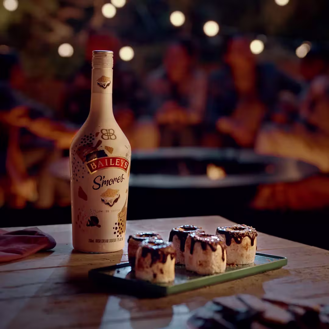 A bottle of Baileys Smores stands on a table next to a plate with four chocolate-drizzled smores; blurred people and string lights are in the background.