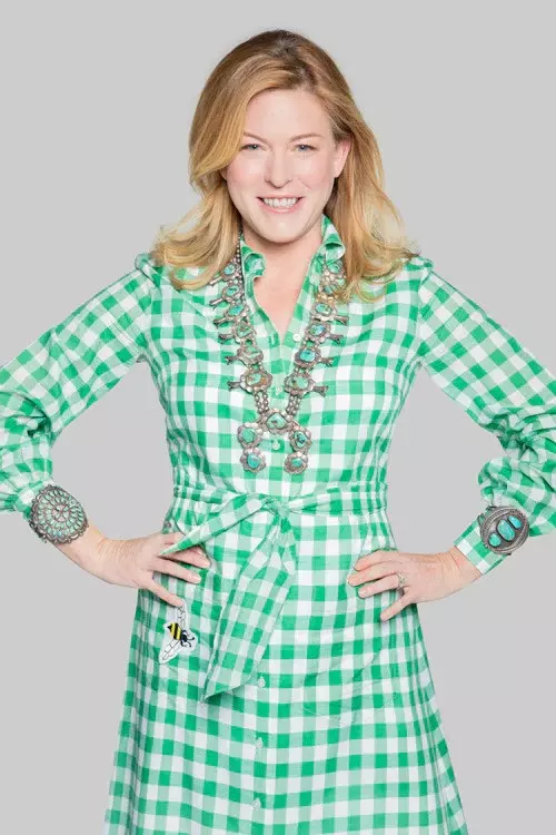 A woman wearing a green and white checkered dress, large statement jewelry, and smiling with her hands on her hips against a plain gray background.
