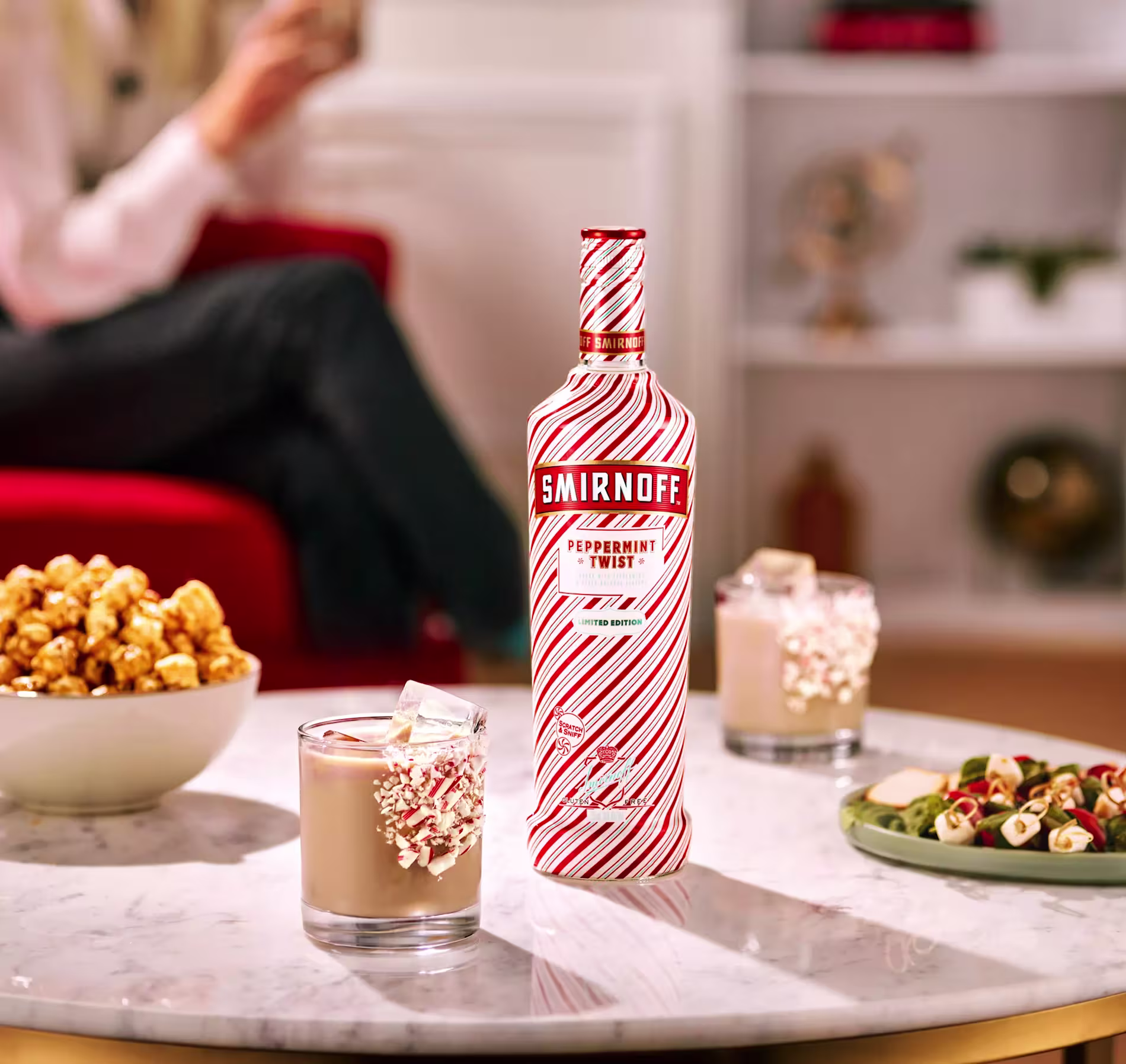 A bottle of Smirnoff Peppermint Twist stands on a marble table with two peppermint-garnished cocktails, caramel popcorn, a snack platter, and blurred people in the background.
