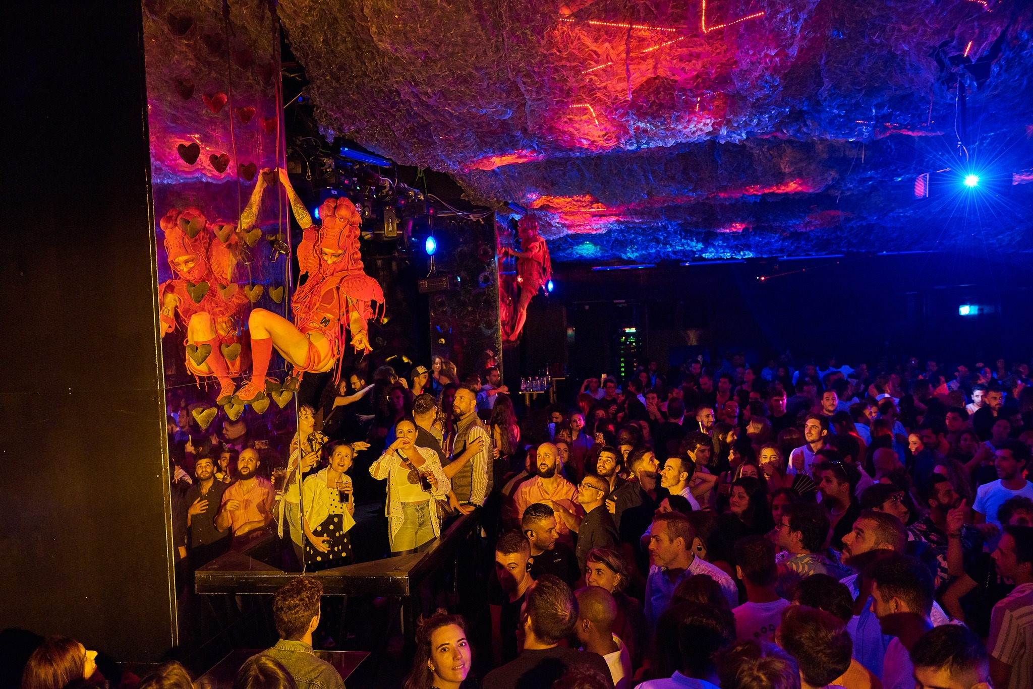 La imagen muestra el Interior de Medias Puri con una multitud de personas, probablemente en un concierto o club. Hay un escenario con artistas, y la iluminación es tenue con algunas luces de colores.