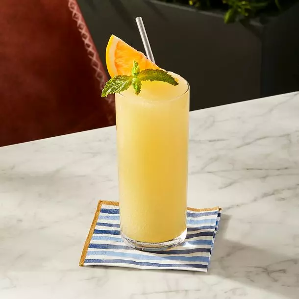 A tall glass of yellow frozen drink with a straw, garnished with an orange slice and mint, sits on a blue-striped napkin on a marble table.