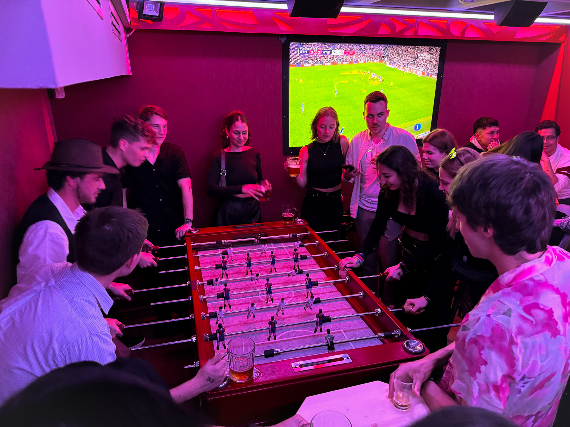 Grupo de personas disfrutando en D9 alrededor de un futbolín, con un partido proyectado en pantalla, creando un ambiente animado y social.