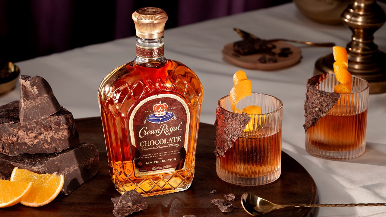 A bottle of Crown Royal Chocolate whisky sits on a tray with two cocktails garnished with orange peel and chocolate pieces, next to chocolate chunks and orange slices.