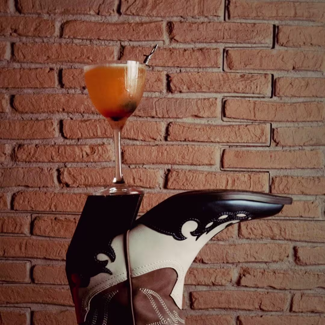 La imagen muestra un cóctel colocado sobre una bota de cowboy de cuero, con detalles en blanco y marrón, sobre un fondo de ladrillos. La bebida tiene una tonalidad dorada y una guarnición en el borde del vaso.