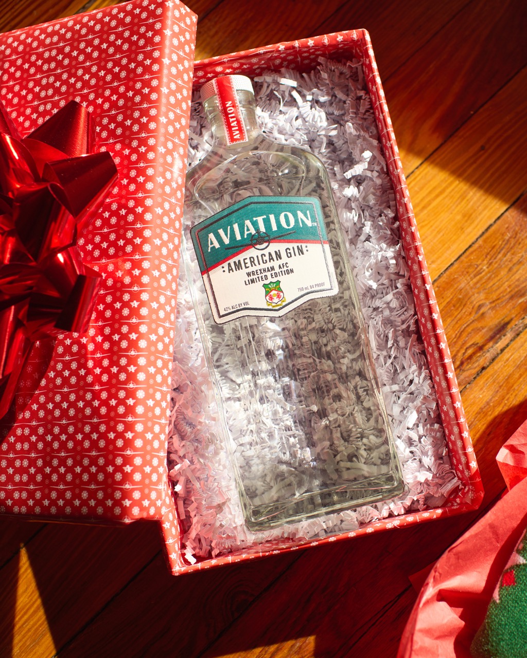 A bottle of Aviation American Gin in a red gift box with white shredded paper, placed on a wooden floor.