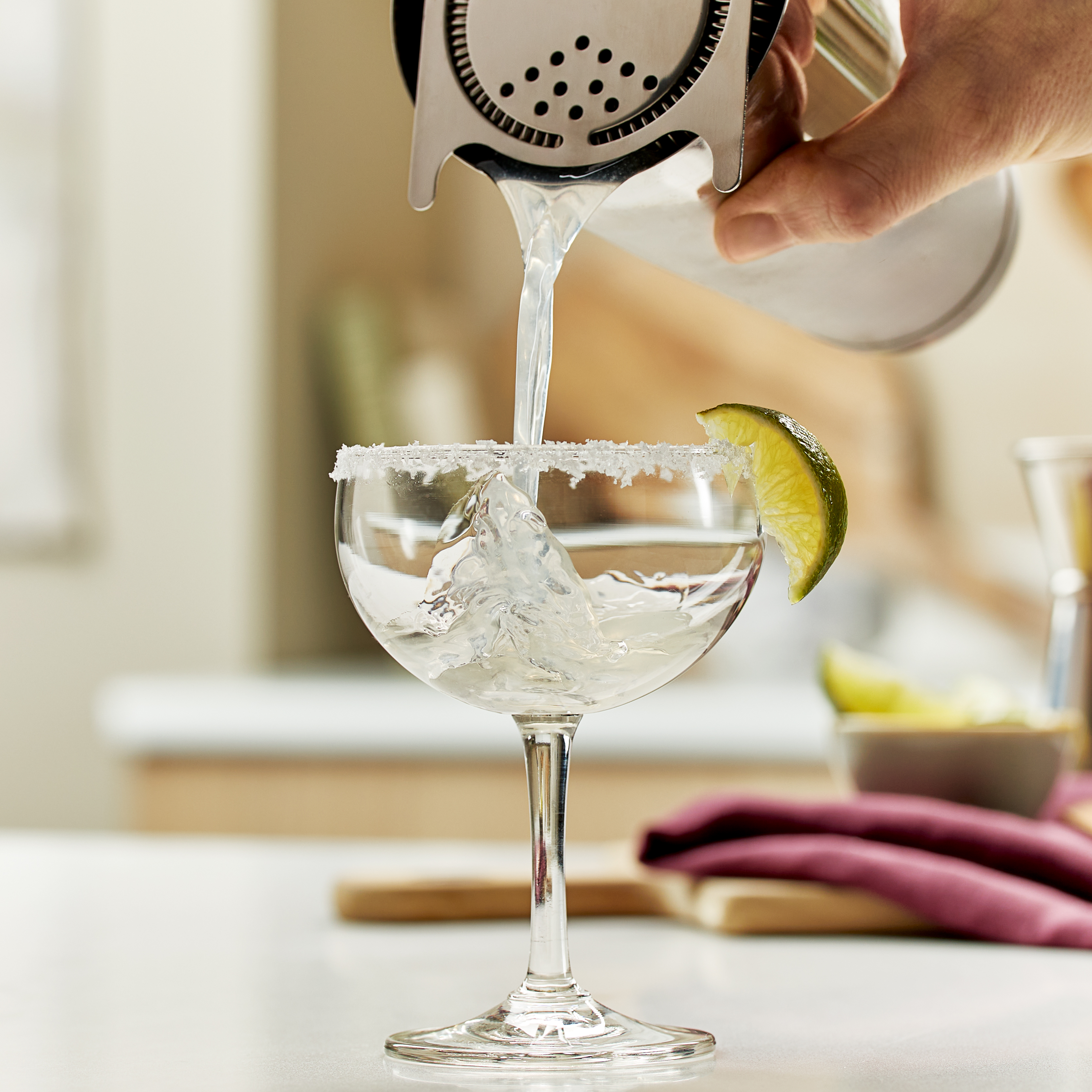 A margarita cocktail is poured and strained from a cocktail shaker into a coupe glass which has been prepared with a salt rim and a lime wedge garnish
