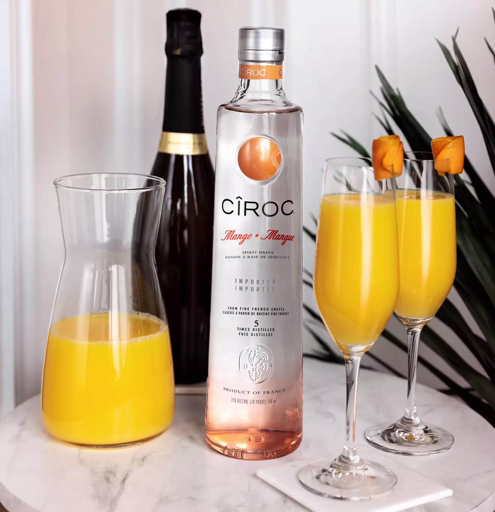 A bottle of Ciroc Mango vodka, a carafe of orange juice, a champagne bottle, and two glasses of orange cocktail garnished with mango cubes are arranged on a white surface.