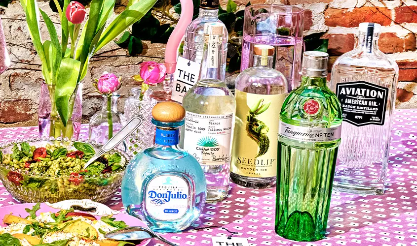 Various bottles of spirits and mixers, including gin, tequila, and non-alcoholic options, are arranged on a pink floral tablecloth with flowers in the background.