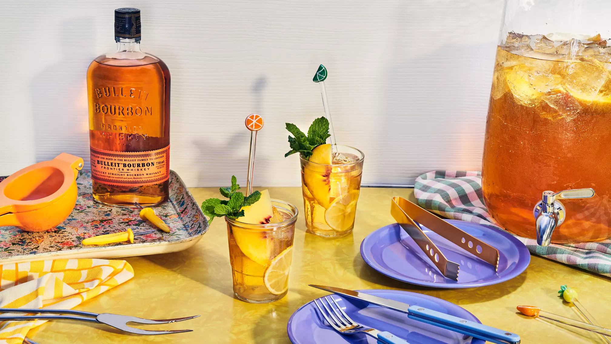 A bottle of Bulleit Bourbon, a pitcher of iced tea, two glasses with lemon and mint, utensils, and a citrus squeezer on a yellow table with a checkered cloth.