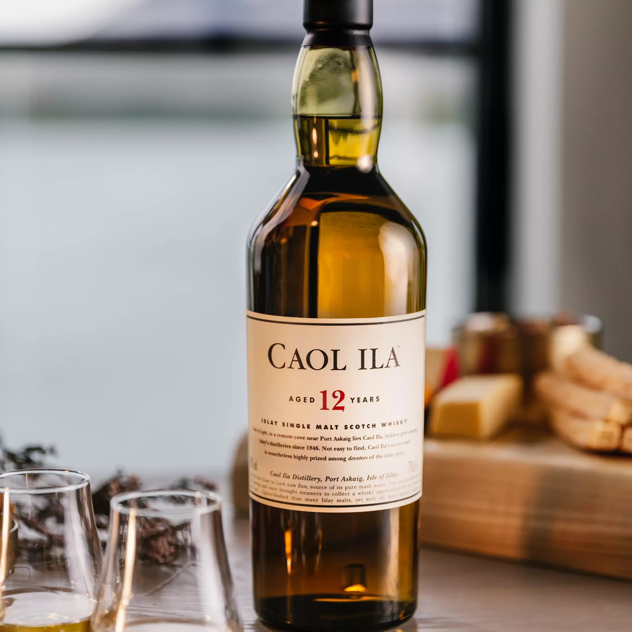 A bottle of Caol Ila 12 Year Old single malt Scotch whisky on a table with two whisky glasses and a cheese board in the background.