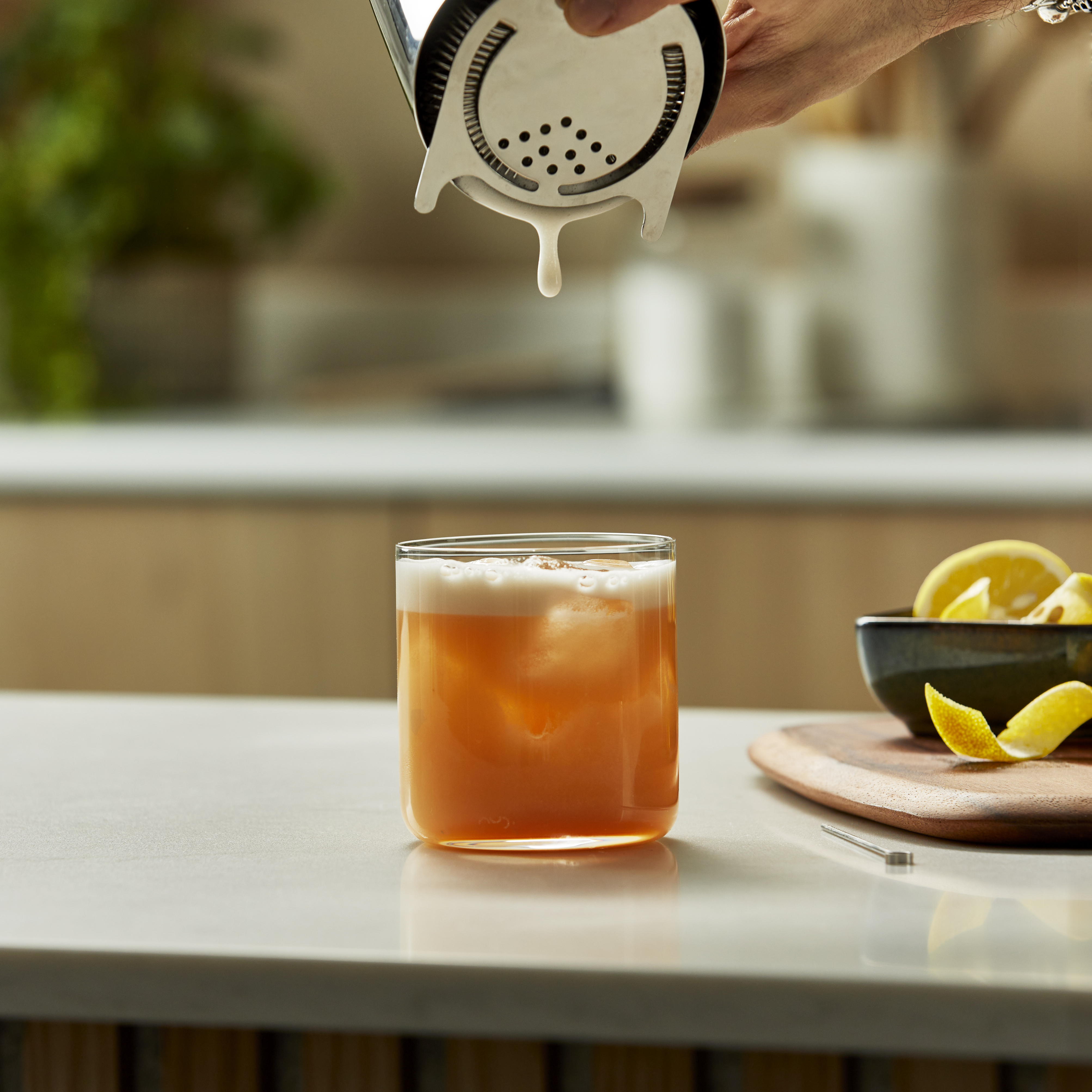 A Whiskey Sour mixture is poured from a cocktail shaker into a glass