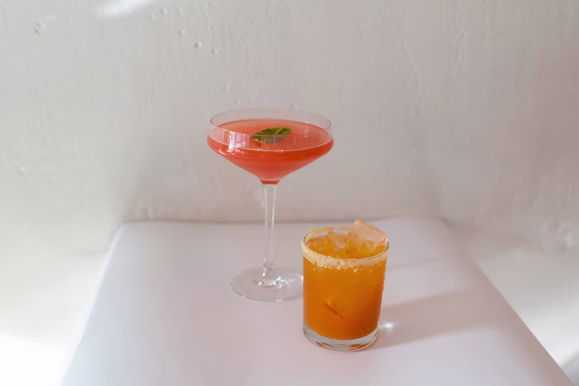 A pink cocktail in a coupe glass with a mint garnish and an orange drink with ice and a salted rim in a short glass on a white surface.