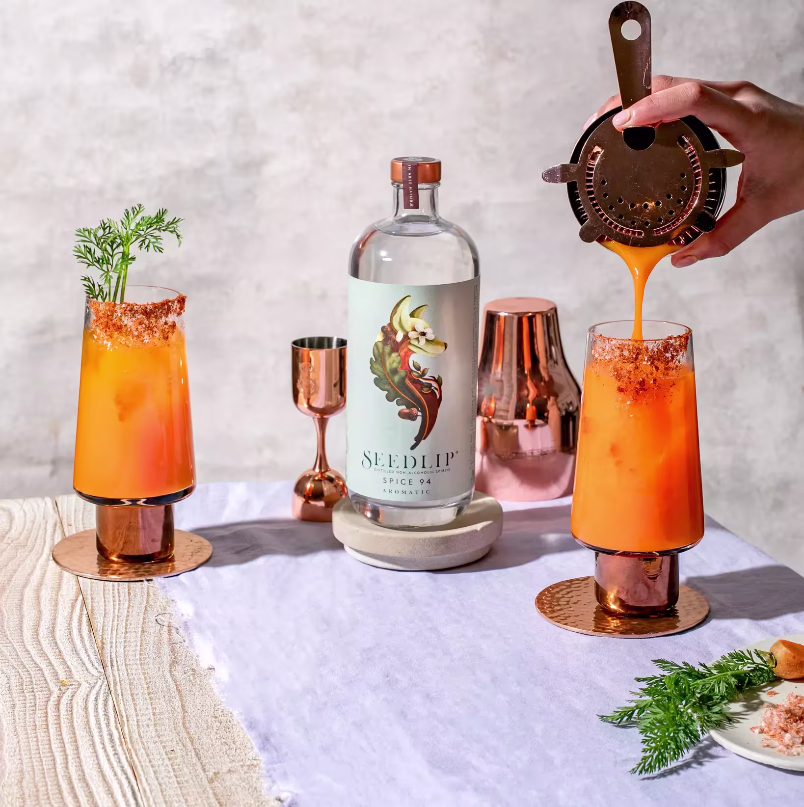 Two orange cocktails in copper glasses with garnish sit beside a Seedlip Spice 94 bottle, as a hand pours liquid into one glass. Copper bar tools and a napkin are also visible.