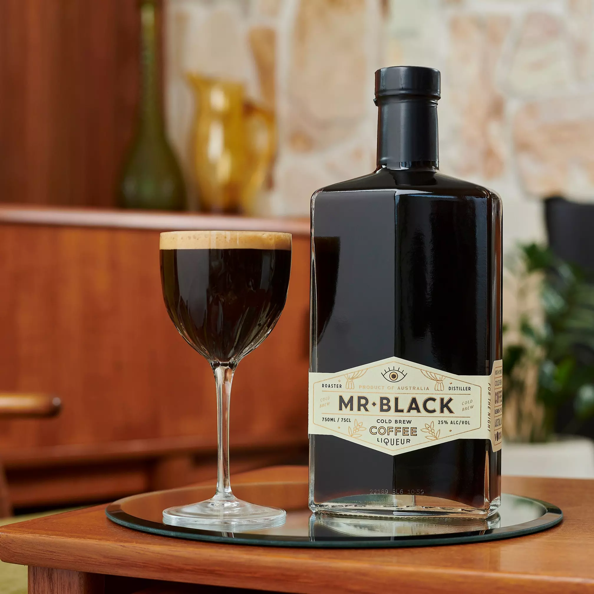 A bottle of Mr Black Cold Brew Coffee Liqueur and a filled cocktail glass sit on a glass tray atop a wooden table in a retro-style room.