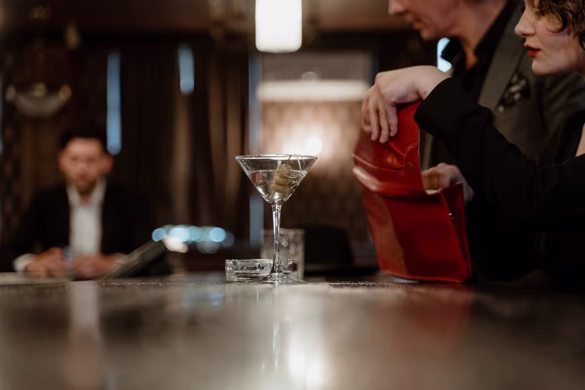 A martini glass with olives and an ashtray on a bar counter; two people with a red clutch bag sit nearby, and another person is in the background.
