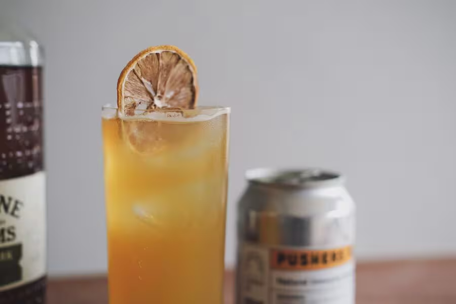 A tall glass filled with an orange drink, ice, and a dried citrus slice garnish, with a can and a bottle partially visible in the background.
