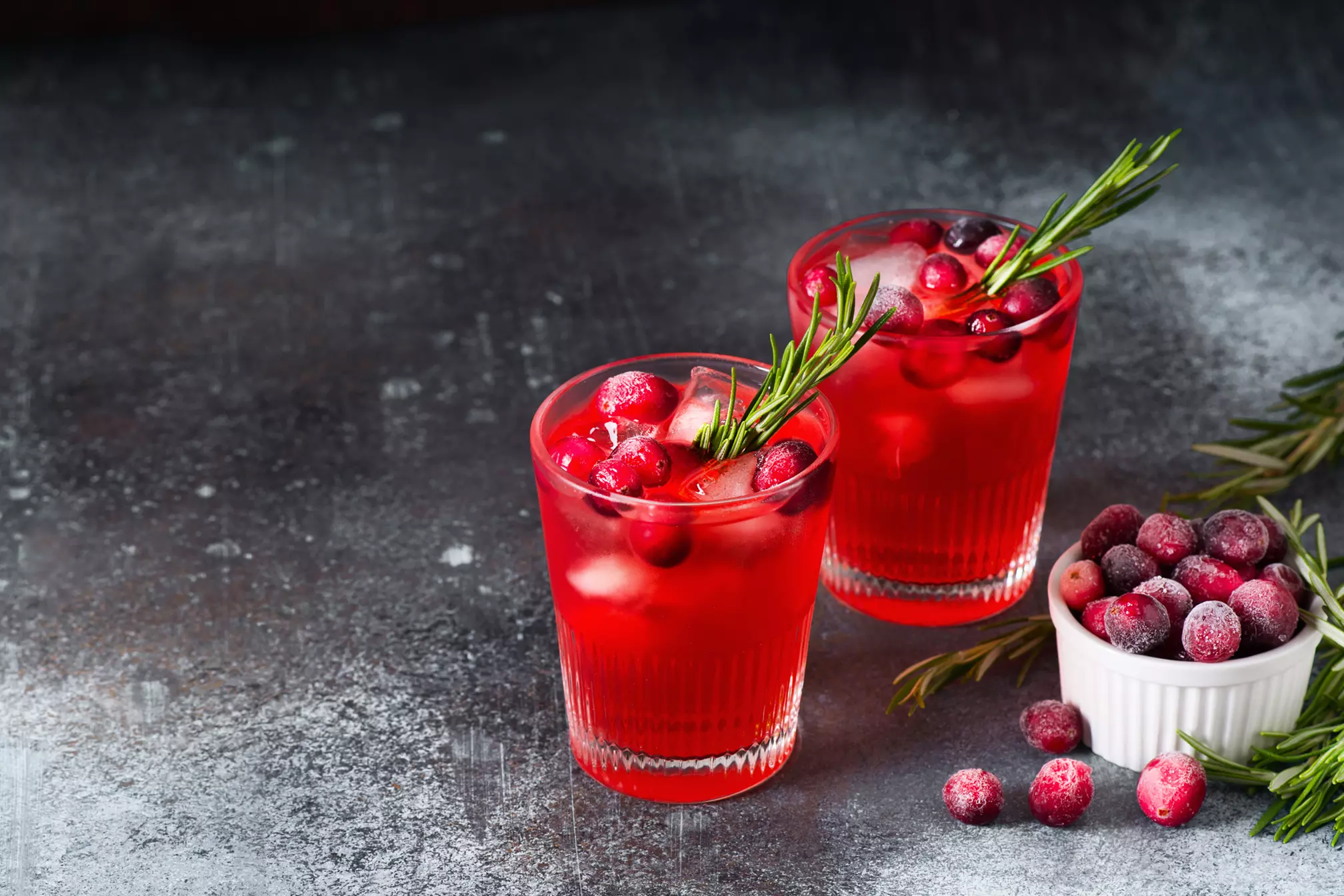 Two glasses of red cranberry cocktail with ice, garnished with rosemary and cranberries, next to a small bowl of frozen cranberries on a dark textured surface.