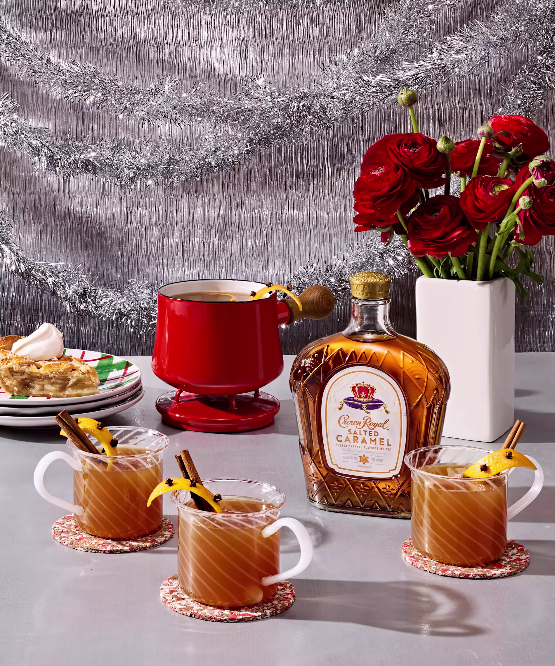 A bottle of Crown Royal Salted Caramel whiskey, three glass mugs of hot drinks with cinnamon sticks, a red fondue pot, pie, and a vase of red flowers on a festive table.