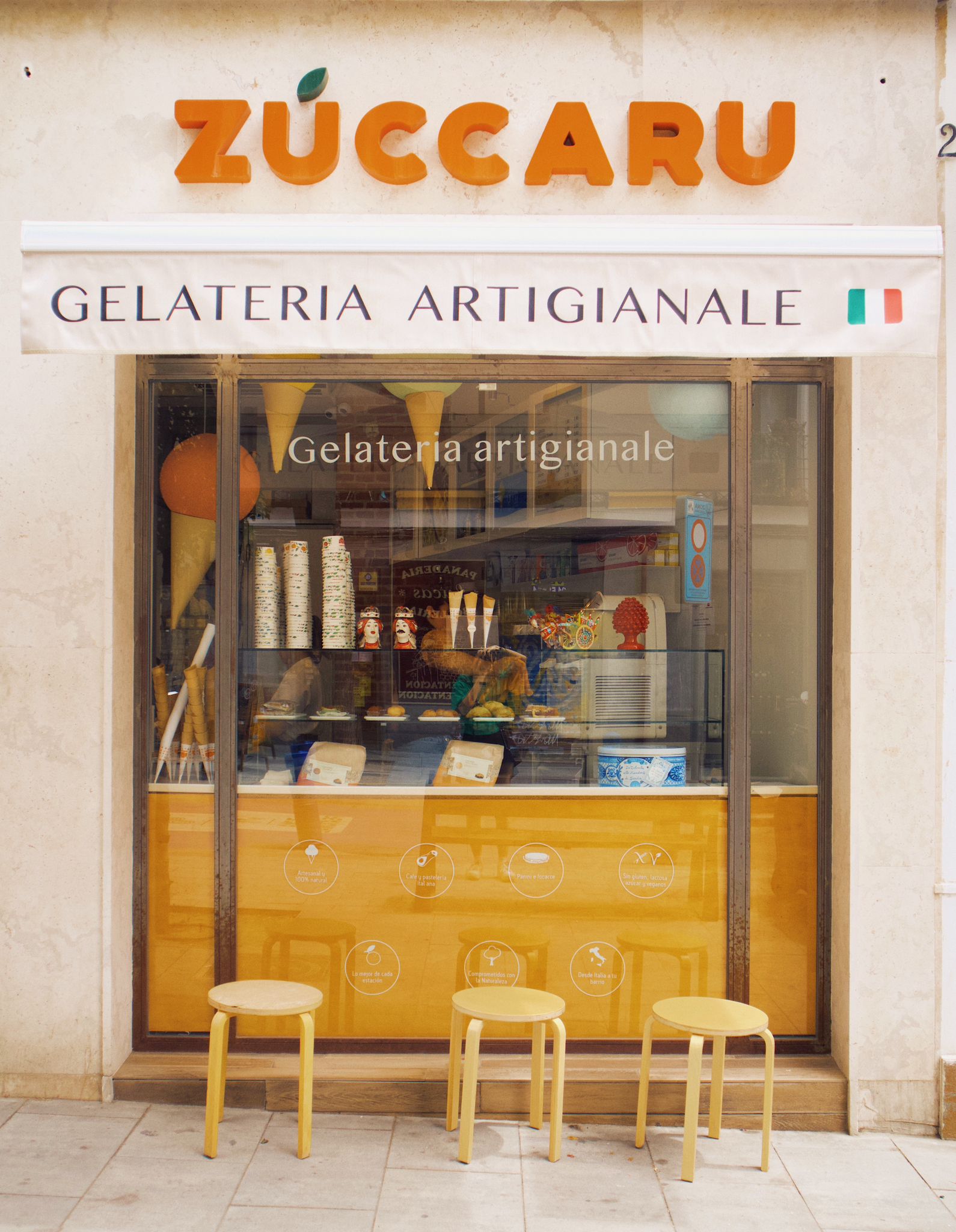 Fachada de la heladería Zùccaru con toldo blanco, letras naranjas, escaparate con conos gigantes y taburetes amarillos frente al ventanal