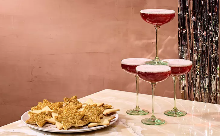 A plate of star-shaped cookies sits next to a pyramid of four coupe glasses filled with a red sparkling drink, set on a marble surface with a pink and silver backdrop.