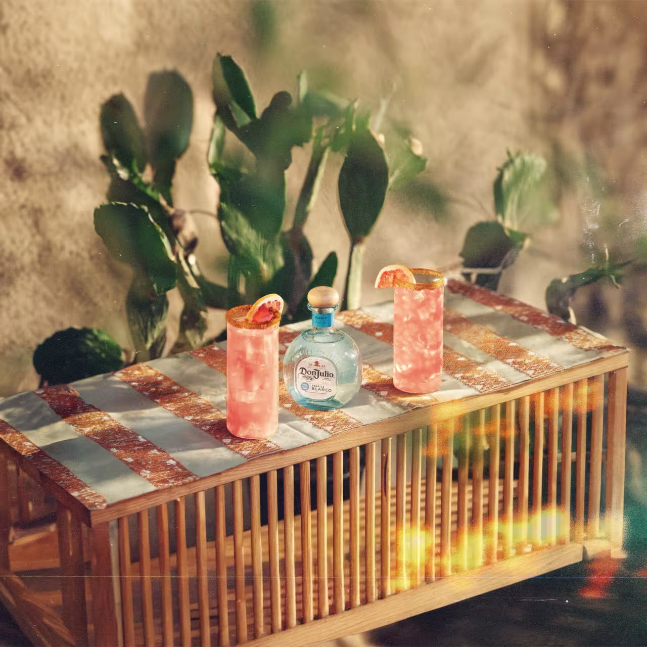 A bottle of Don Julio tequila and two pink cocktails with grapefruit garnishes sit on a striped table, with green plants in the background.