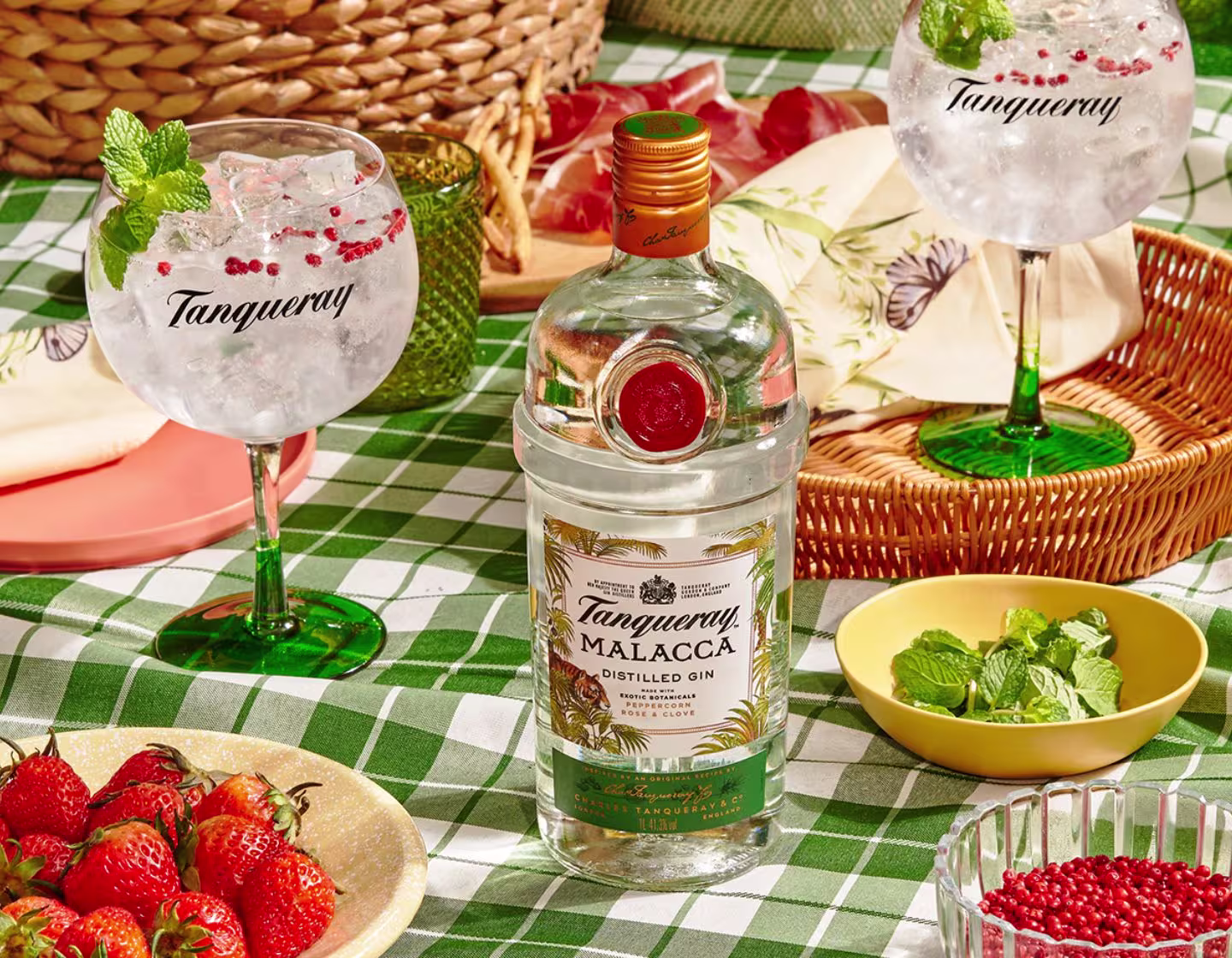 A bottle of Tanqueray Malacca gin on a checkered tablecloth, surrounded by gin cocktails, strawberries, mint leaves, and bowls of garnishes.