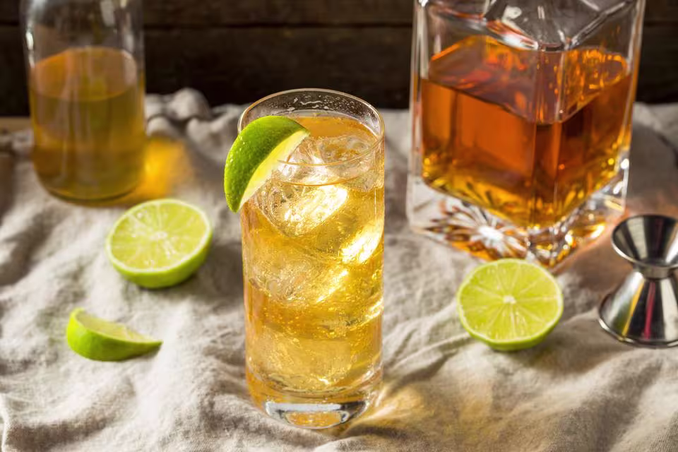 A glass of iced whiskey cocktail garnished with a lime wedge, surrounded by bottles of liquor, lime halves, and a metal jigger on a cloth surface.