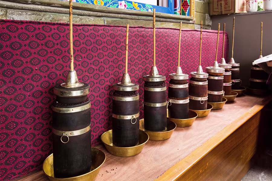 Several traditional wooden Tibetan butter tea churns with metal tops stand in brass bowls on a wooden surface against a red patterned backdrop.