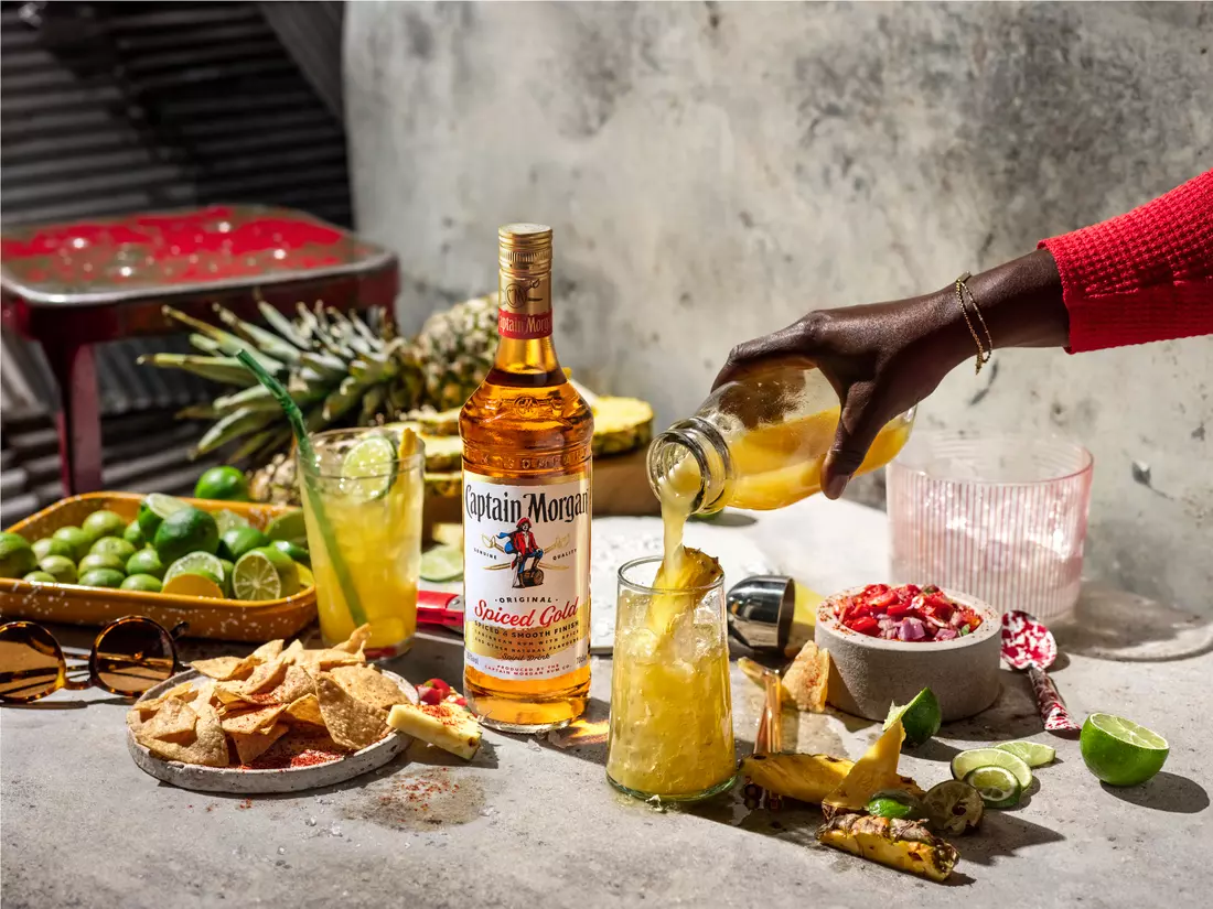 A hand pours a yellow drink into a glass beside a bottle of Captain Morgan Spiced Gold, with chips, fruit, and lime wedges on a concrete table.