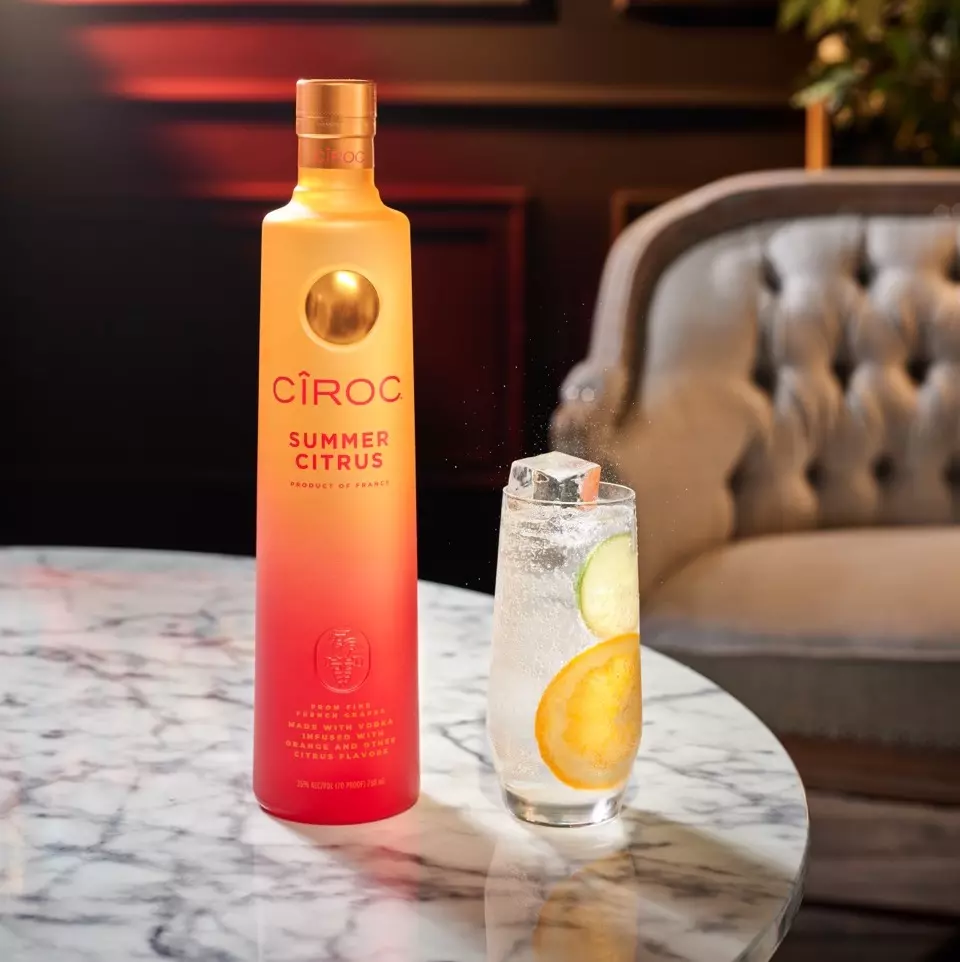 A bottle of Ciroc Summer Citrus vodka stands next to a glass of sparkling beverage with ice, lemon, and cucumber slices on a marble table.
