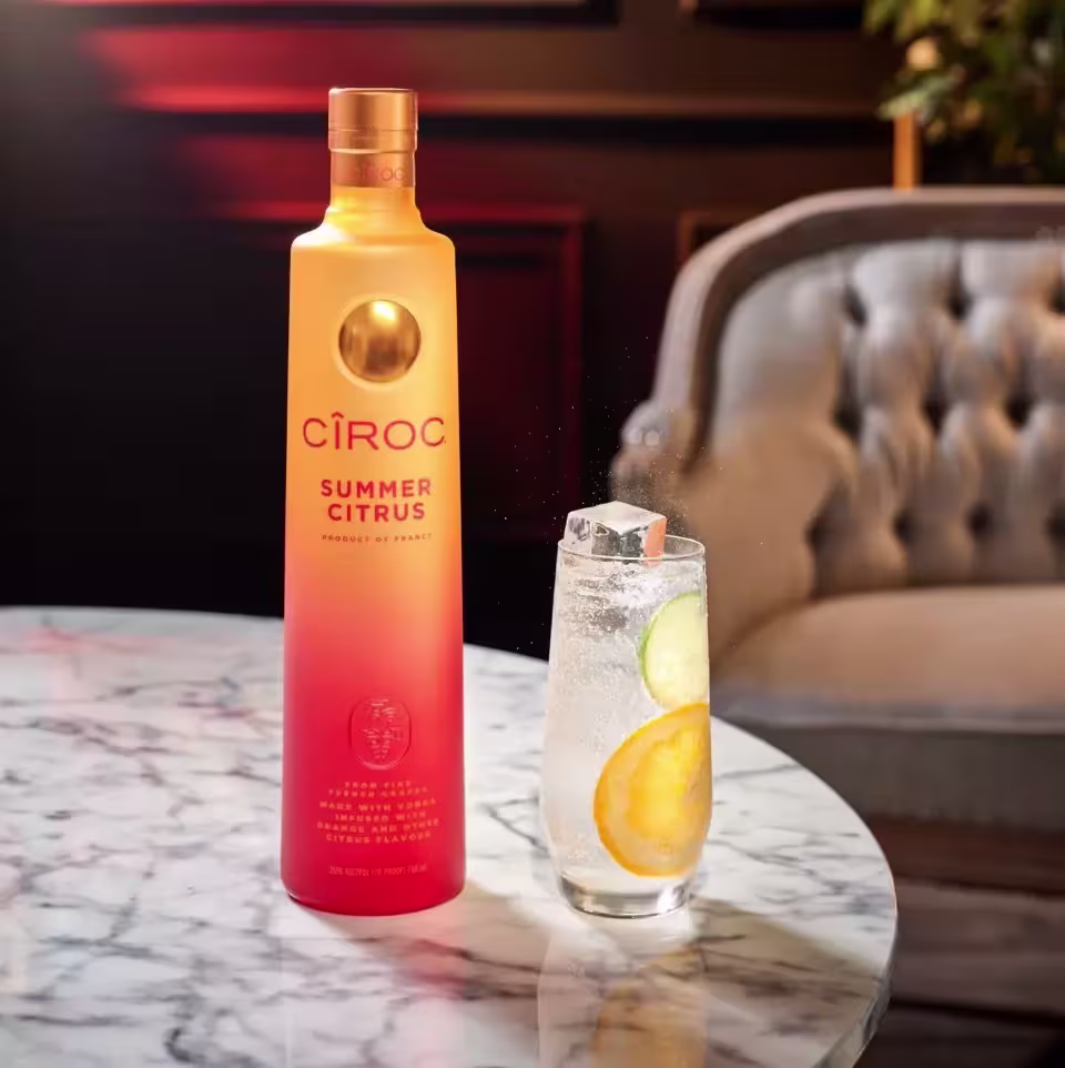 A bottle of Ciroc Summer Citrus vodka stands next to a glass of sparkling beverage with ice, lemon, and cucumber slices on a marble table.