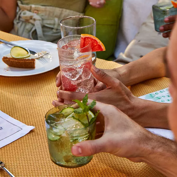 Three people hold different cocktails at an outdoor table, one with a sprig of mint and cucumber, another with a grapefruit slice. A dessert plate with lime garnish is in the background.