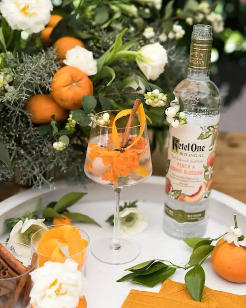 A glass of cocktail with orange slices and a cinnamon stick beside a Ketel One Botanical Peach & Orange Blossom bottle, surrounded by flowers and fresh oranges.