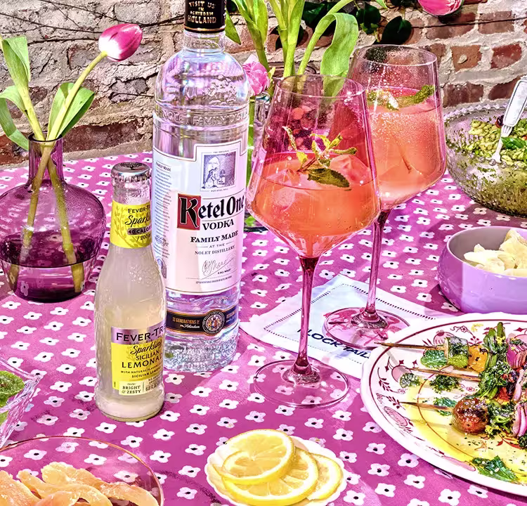 A table set with Ketel One vodka, Fever-Tree Sicilian Lemonade, two pink cocktails, sliced lemons, a floral vase, and plates of food on a pink patterned tablecloth.