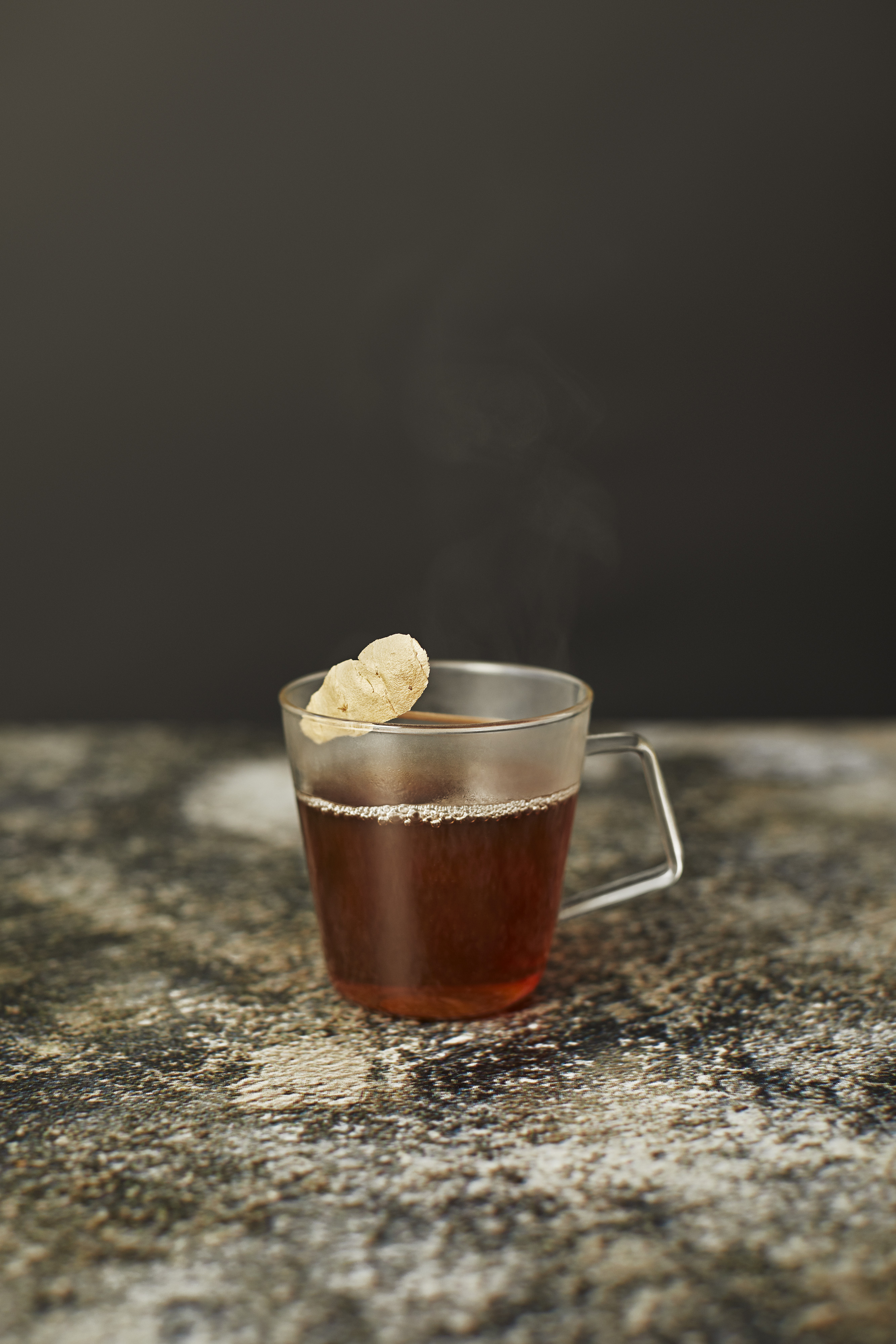 A clear glass cup filled with dark tea, topped with a slice of ginger, sits on a textured surface against a dark background.