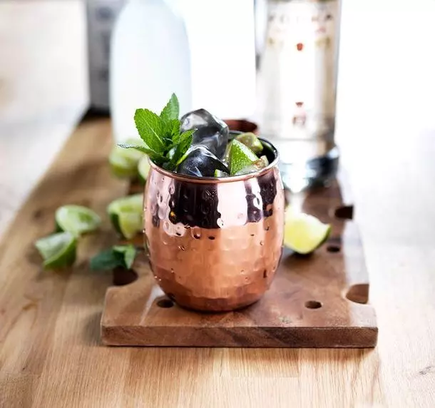 A copper mug filled with ice and mint sits on a wooden board, surrounded by lime wedges and bottles in the background.