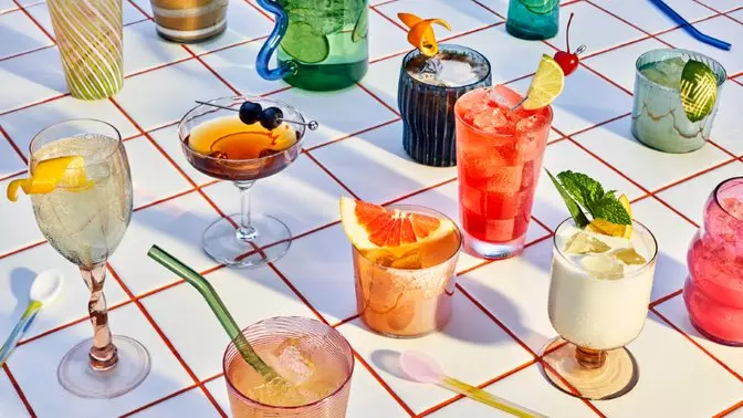 An assortment of colorful cocktails and drinks in various glasses arranged on a white and red tiled surface, with straws, garnishes, and spoons.
