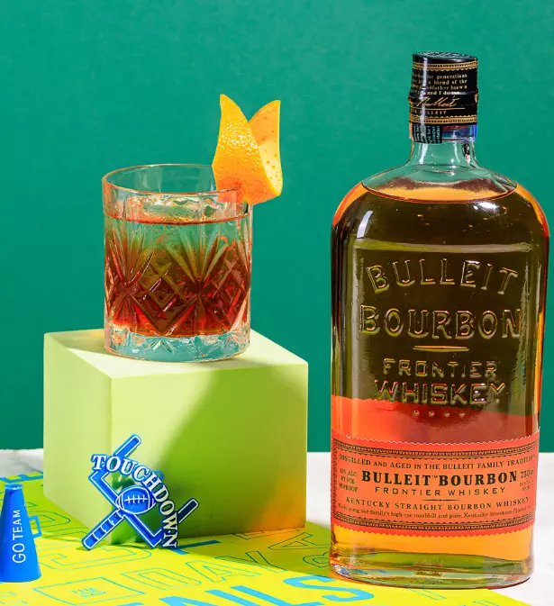 A bottle of Bulleit Bourbon and a glass with an orange peel garnish are displayed on geometric props with a football-themed coaster in a colorful setting.